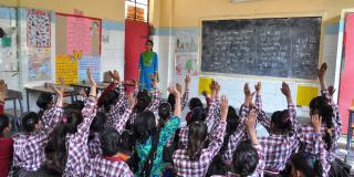 Girls sit on the floor of their classroom and raise their hands; at the front, a young female teacher stands by the blackboard