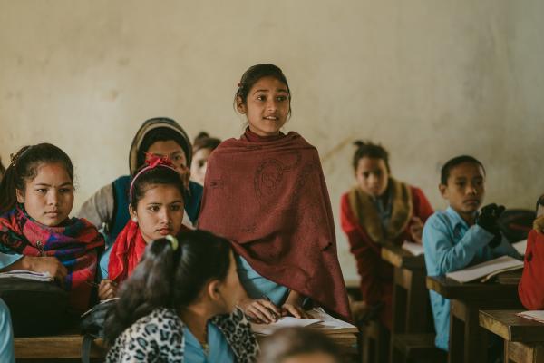 A girl stands up to speak in the classroom, surrounded by her classmates sitting at their desks.