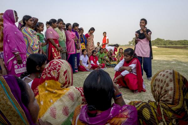 Women sit and stand in a semi-circle to watch two young women perform an interactive play about child marriage in Dobini, Nepal.