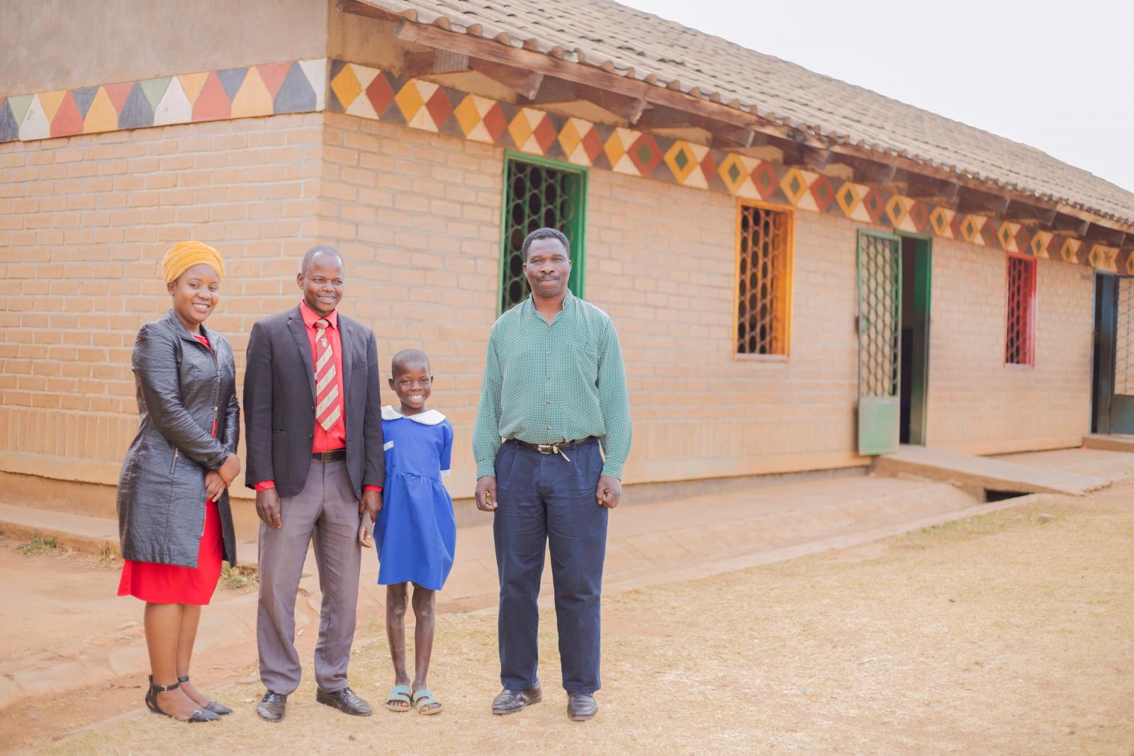 VSO volunteer Prisca, teacher Peter, student Alinafe, and VSO volunteer Theo, standing outside a classroom block.