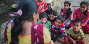 A group of young children sit outside on the round, listening to a local teacher talk about school and earthquake preparedness