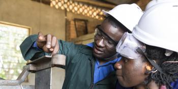 A student watches intently as her instructor demonstrates machinery in a welding and metal workshop