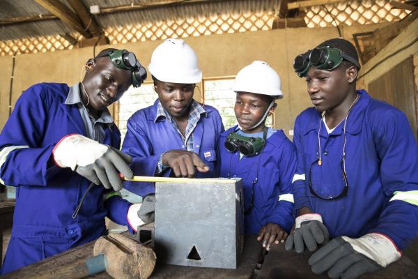 Four students dressed in overalls and safety wear gather around a metal box in a metalwork worshop