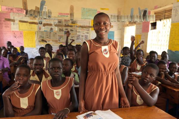 A girl in school uniform smiles as she stands up at her desk, surrounded by classmates who are sitting down