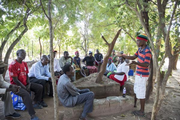 Former peer educator JK stands and speaks animatedly to a group of people who are sitting outside and listening to him